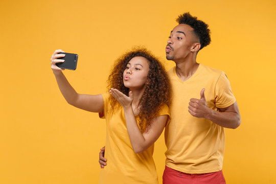 Pretty friends couple african american guy girl in casual clothes isolated on yellow background. People lifestyle concept. Mock up copy space. Doing selfie shot on mobile phone blowing send air kiss.