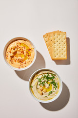 Top view of bowls with delicious hummus and crackers on grey background