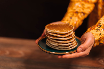 Female hands in a yellow dress hold out a saucer with pancakes with honey.
