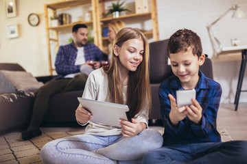 Children sister and brother playing using phone and digital tablet on floor while young parents relaxing at home on sofa.