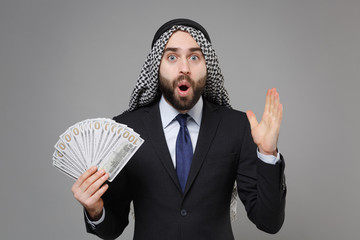 Shocked bearded arabian muslim businessman in keffiyeh kafiya ring igal agal suit isolated on gray background. Achievement career wealth business concept. Hold fan of cash money in dollar banknotes.