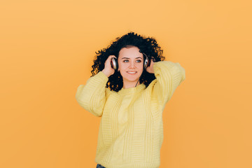 young beautiful girl with curly hair listens to music on headphones in a yellow sweater on a yellow background