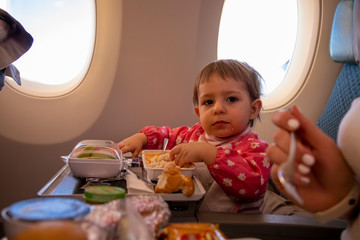  young toddler passenger plane eats tasty hot special  meal  on board  on a folding table. in the background is a window in the porthole.