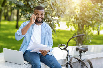 Smiling Black Businessman With Papers And Laptop Talking On Cellphone Outdoors
