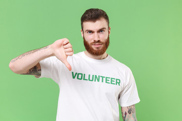 Perplexed young bearded man in white volunteer t-shirt isolated on pastel green wall background...