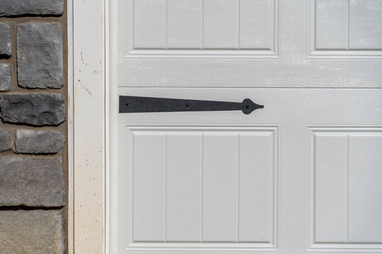 Black Powder Coated Steel Spade Strap Hinge Pointing To Left On A Raised Panel White Garage Door Attached To Ledge Stone Veneer Covered Wall  On An American Single Family House