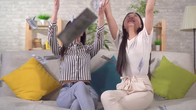 two happy asian girls catch flying dolar banknotes while sitting at home on the sofa in the living room