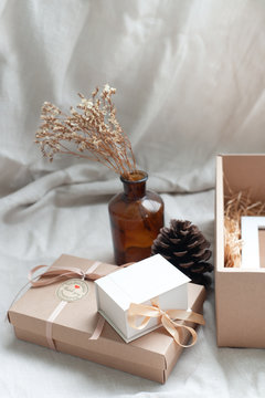 A white picture frame placed in a brown gift box surrounded by a box of small diamond rings tied with cream-colored pine cones dried flowers in a brown glass bottle. Put on a light brown fabric