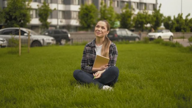 long shot. 20 years old girl in a plaid shirt sits on the grass and laughs sincerely 4K