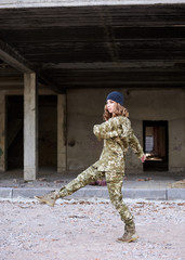 Young curly blond military woman, wearing ukrainian military uniform and dark grey hat, marching. Full-length portrait of army soldier walking in front of ruined abandoned building, construction site.