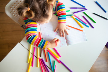 Little girl, a 3 year old girl, with a ponytail hairstyle in a multi-colored colorful striped jacket on a light background at the table draws multicolored markers and smiles