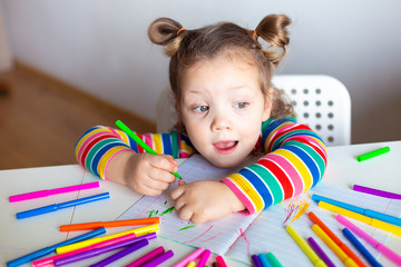 Little girl, a 3 year old girl, with a ponytail hairstyle in a multi-colored colorful striped jacket on a light background at the table draws multicolored markers and smiles
