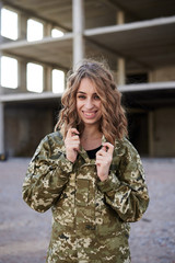 Young curly blond military woman, wearing ukrainian army military uniform and black t-shirt, posing. Three-quarter portrait of female soldier in front of ruined abandoned building,construction site.