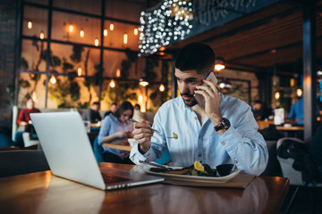man using mobile phone and laptop in restaurant while having lunch