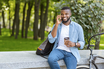Obraz premium Handsome Afro Guy Sitting In Park With Coffee, Talking On Cellphone