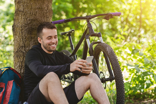 Smiling guy sitting next to bike, chatting on smartphone