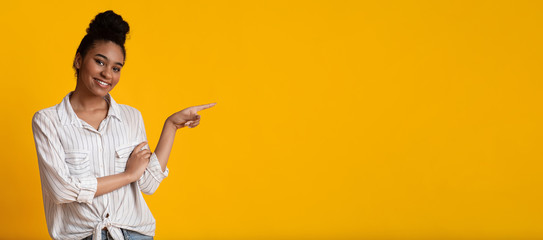 Millennial afro girl pointing aside at copy space on yellow background