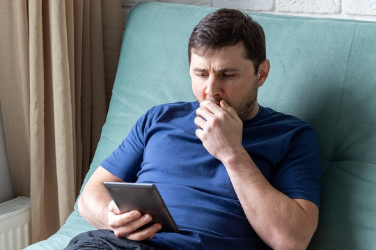 Man Yawns While Reading An E-book. A Resting Male In A Blue T-shirt Sits On A Green Sofa With An Electronic Book In One Hand, Yawns, Covering His Mouth With His Second Hand