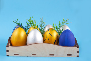 Christ is risen. Easter eggs painted in silver, gold and blue lie in a wooden box with sprigs of fresh herbs. Classic blue background.
