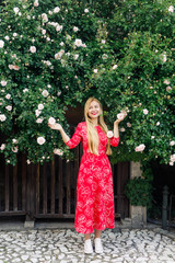 portrait of a young stylishly dressed girl in a red dress standing in the garden near a rose bush