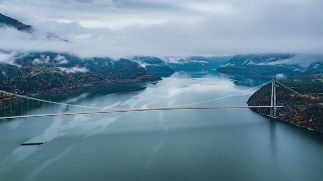 The suspension bridge spanning above the calm blue water of the Eidfjorden branch of Hardangerfjorden, connecting Ullensvang and Ulvik regions. Thick white clouds hanging above.