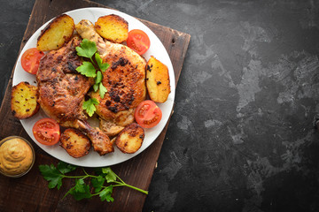 Fried chicken thigh, baked chicken leg. Top view. Dark background. Plate on a wooden board. Baked potatoes, tomatoes and chicken, a delicious lunch.
