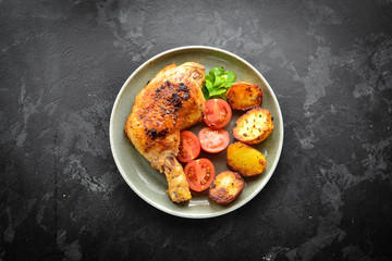 Fried chicken thigh, baked chicken leg. Top view. Dark background. Plate on a wooden board. Isolated. Baked potatoes, tomatoes and chicken, delicious lunch.