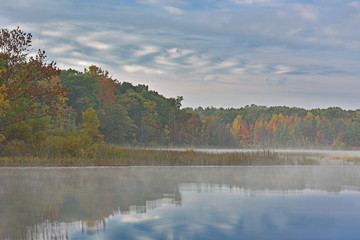 Foggy autumn landscape at dawn of the shoreline of Deep Lake with mirrored reflections in calm water, Yankee Springs State Park, Michigan, USA
