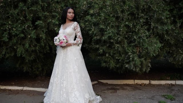 attractive African american lady in fashionable wedding dress holds rose flowers near green bushes slow motion