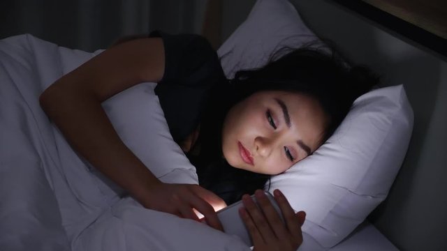 Asian young woman lying on bed play smartphones during night time In bedroom that turns off lights. Read COVID-19 on mobile phone. Using p hone in low light has health effect on eyes. Insomnia Concept