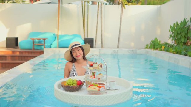 Young asian woman in swimming pool with breakfast from floating basket. Luxurious breakfast in resort