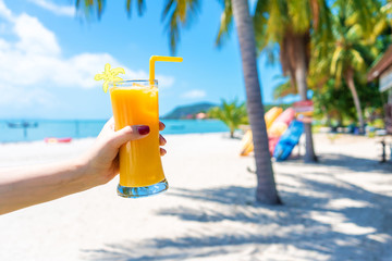 First-person view. Girl holds a glass cup of cold mango fresh on the background of a sandy tropical beach. White sand and palm trees. Fairytale vacation