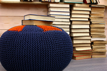 A stack of books,   lots of books on the floor and on a bookshelf. Knitted soft pouf on the...