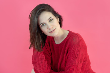 Fototapeta premium Portrait of an attractive brunette girl with green eyes in a red sweater on a pink background. Studio photo
