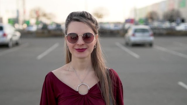 Portrait of a Woman in Sunglasses and a Maroon Dress Walking in The Parking lot of a Shopping Center. Close up