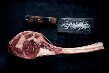Raw Tomahawk steak dry aged with a meat cleaver on a dark stone background, top view
