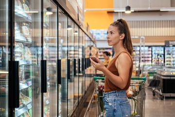 Woman using mobile phone at grocery store stock photo. Woman standing in frozen section of grocery store with cart and smartphone