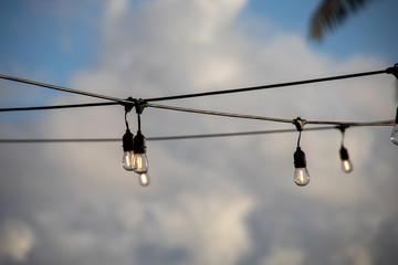 A string of outdoor lighting with on and off light bulbs hanging in the air, against a bright blue sky with clouds. Early morning feeling, selective focus on light bulbs.