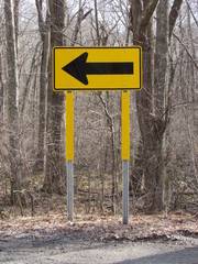 View of a yellow direction traffic sign on the side of a country road with woods in the background 