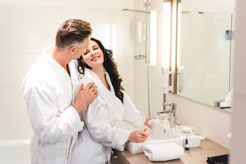 boyfriend hugging smiling girlfriend in bathrobe with toothbrush in hotel