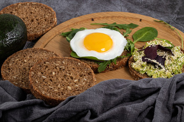 Avocado sandwich on dark rye bread with spinach and fried egg on a dark background