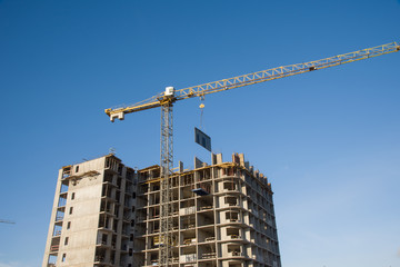 Tower crane lifts the block for building under construction at background blue sky at sunny day. Precast concrete slab hanging from crane hook above building skeleton at construction site