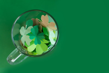 Top view of bear transparent cup with different shades of green four-leafed paper shamrocks on green background