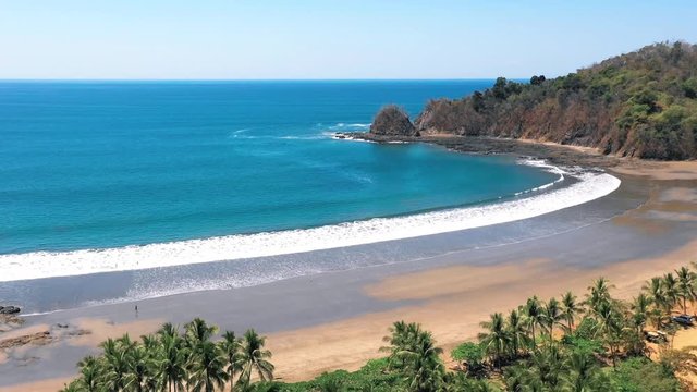 Long pull back aerial reveal over idyllic palm tree tropics, Costa Rica sandy beach and ocean waves