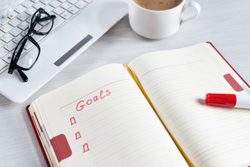 List with goals in notebook, computer and cup of coffee on white wooden table.