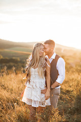 Beautiful couple in love embracing, at the sunset in summer field. Girl in white summer dress and feathers in hair, man in stylish casual suit. Summer vacation concept, boho couple