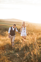 Fototapeta na wymiar Handsome stylish man in rustic suit and pretty boho woman in dress walking in the field with straw bales, holding hands and smiling. Summer evening, sunset
