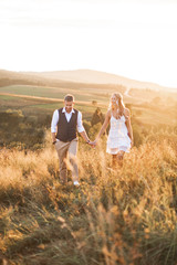 Handsome stylish man in rustic suit and pretty boho woman in dress walking in the field with straw bales, holding hands and smiling. Summer evening, sunset