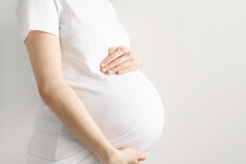 Pregnant woman in a white t-shirt holds her belly with her hands on a light background with copyspace holds her belly with hands. Motherhood and love