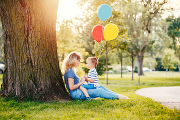 Young Caucasian mother and boy toddler son sitting on green grass together in park. Family mom and child talking communicating outdoor on a summer day. Happy authentic family childhood lifestyle.